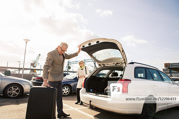 Senior man loading luggage in car trunk with woman standing in parking lot against sky