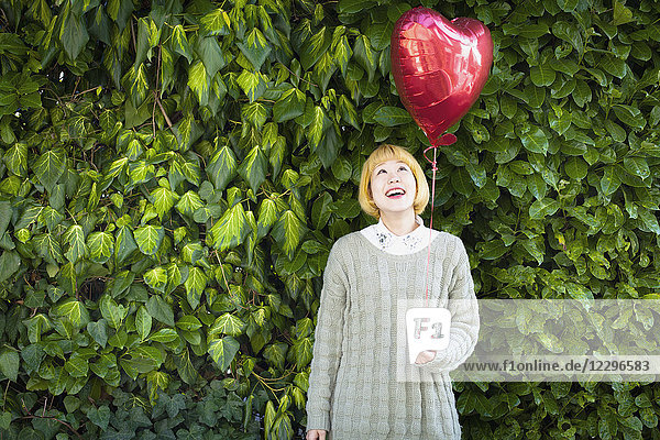 Lächelnde junge Frau hält einen Ballon in Herzform  während sie gegen Pflanzen steht.