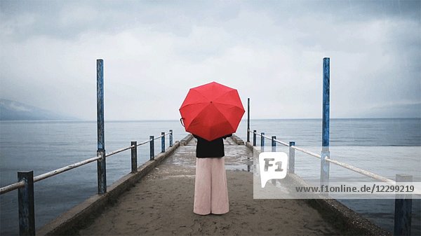 Rückansicht einer Frau  die an einem regnerischen Tag einen roten Regenschirm am Pier dreht