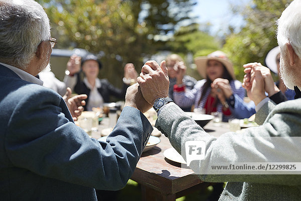 Active senior friends holding hands  praying at sunny garden party table