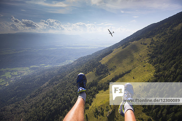 Die Füße eines Gleitschirmfliegers baumeln hoch über den slowenischen Alpen bei einem Gleitschirmflug vom Berg Dobrca bei Begunje und Radovljica in Slowenien. Im Hintergrund ein Segelflugzeug.