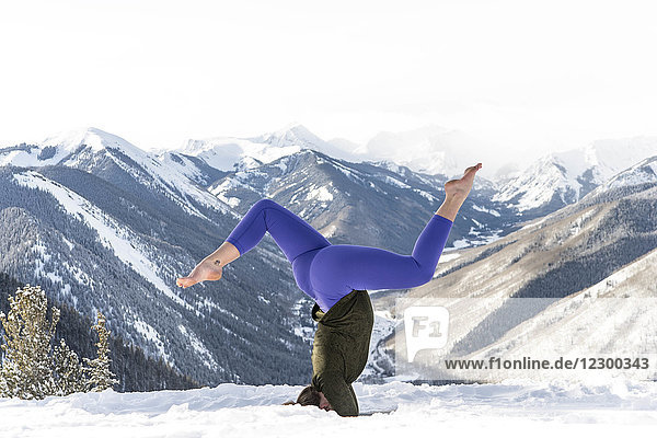 Junge Frau macht Kopfstand auf einem verschneiten Hügel vor einem Bergtal  Aspen  Colorado  USA