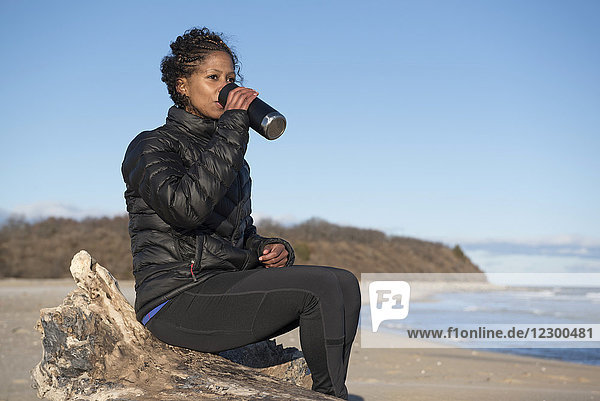 Frau mit schwarzen lockigen Haaren trinkt am Strand Kaffee aus einer Thermoskanne
