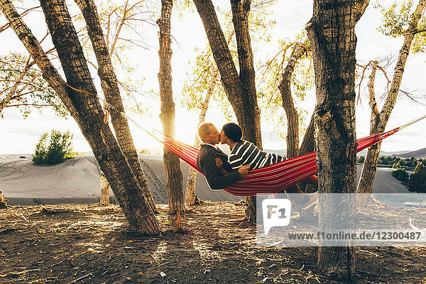 Ein junges Paar küsst sich in einer Hängematte in den Sanddünen  während die Sonne im Hintergrund untergeht.