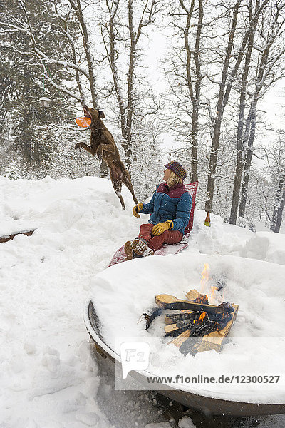 Frau entspannt sich im Schnee an einer brennenden Feuerstelle und spielt mit einem Hund Plastikring  Durango  Colorado  USA