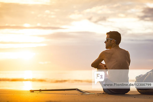 Rückansicht eines einsamen Mannes ohne Hemd  der bei Sonnenaufgang am Strand sitzt