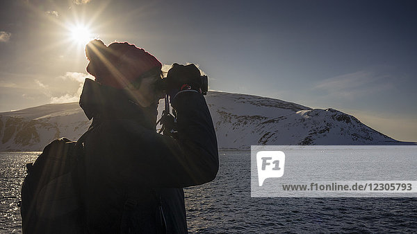 Frau mit Rucksack schaut durch ein Fernglas in arktischer Landschaft mit Meer und Bergen im Hintergrund  Spitzbergen  Svalbard und Jan Mayen  Norwegen