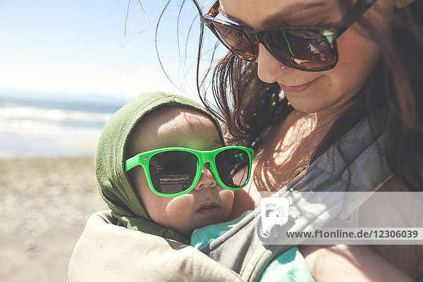 Mutter hält ihren kleinen Sohn am Strand und beide tragen eine Sonnenbrille