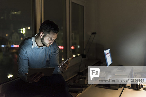Businessman sitting on windowsill in office at night looking at smartphone