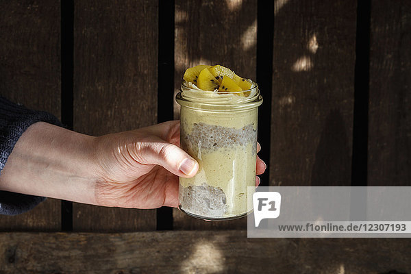 Man's hand holding glass of chia pudding with mango smoothie garnished with kiwi