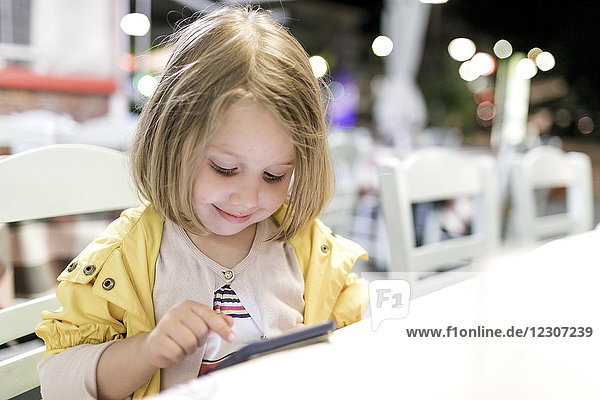 Porträt eines lächelnden kleinen Mädchens,  das in einem Restaurant sitzt und mit dem Smartphone spielt.