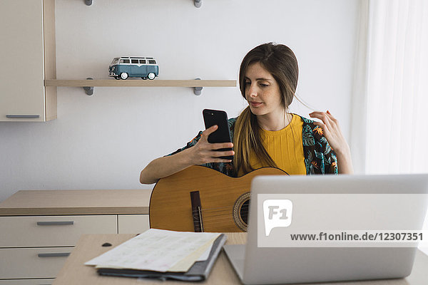 Young woman sitting at table at home with guitar  cell phone and laptop