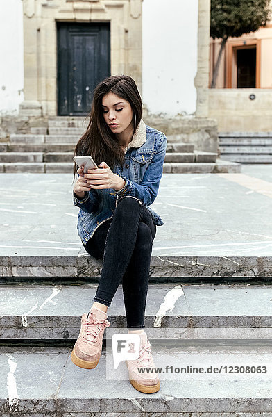 Junge Frau sitzt auf einer Treppe in einer Stadt und überprüft ihr Smartphone.