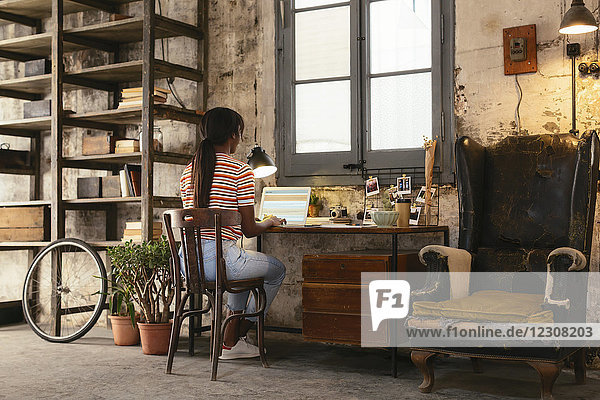 Back view of young woman sitting at desk in a loft working on laptop