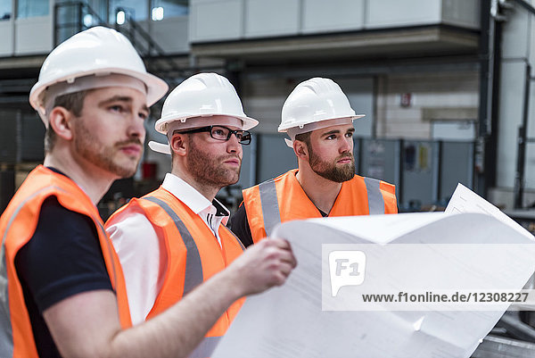 Three men wearing hard hats and safety vests holding plan in factory