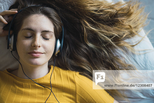 Porträt einer lächelnden jungen Frau  die auf dem Bett liegt und mit Kopfhörern Musik hört.