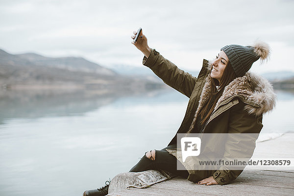 Lächelnde junge Frau sitzt auf dem Steg und nimmt Selfie mit Smartphone