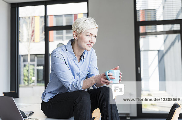 Lächelnde Frau mit Tasse Kaffee im Büro auf dem Tisch sitzend