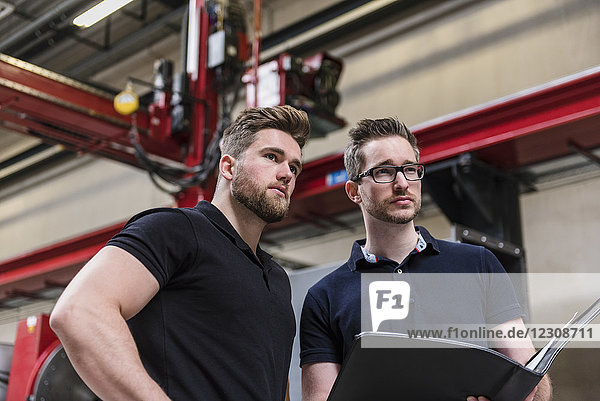 Two men with folder on factory shop floor looking around