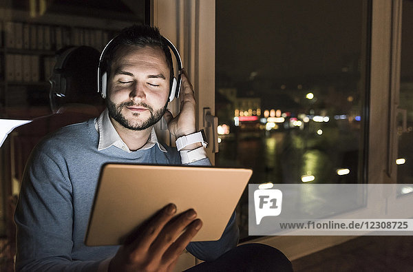 Geschäftsmann sitzt nachts auf der Fensterbank im Büro und hört Musik mit Kopfhörer und Tablett.