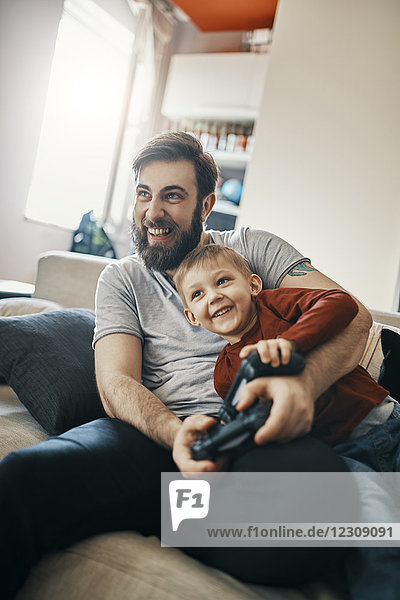 Lachender Vater und kleiner Sohn sitzen zusammen auf der Couch und spielen ein Computerspiel.