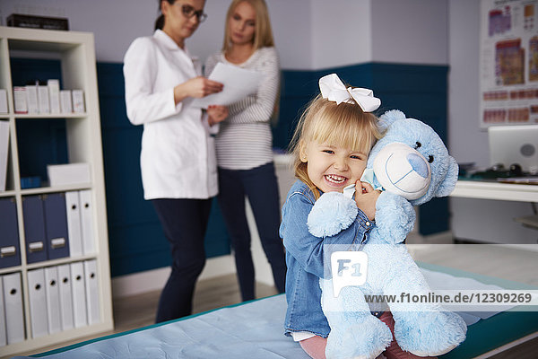 Mädchen umarmender Teddy in der Arztpraxis mit Arzt und Mutter im Hintergrund
