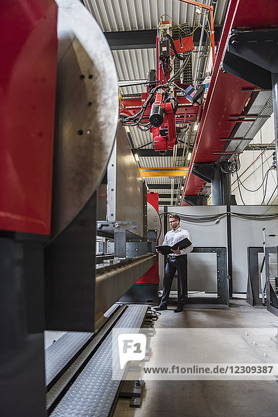 Businessman with folder standing at machine on factory shop floor