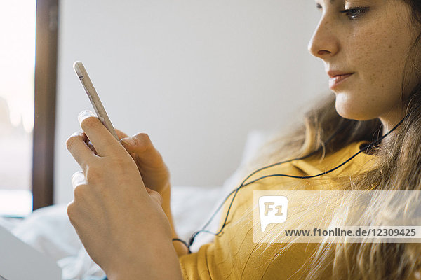 Junge Frau auf dem Bett liegend mit Handy und Kopfhörer