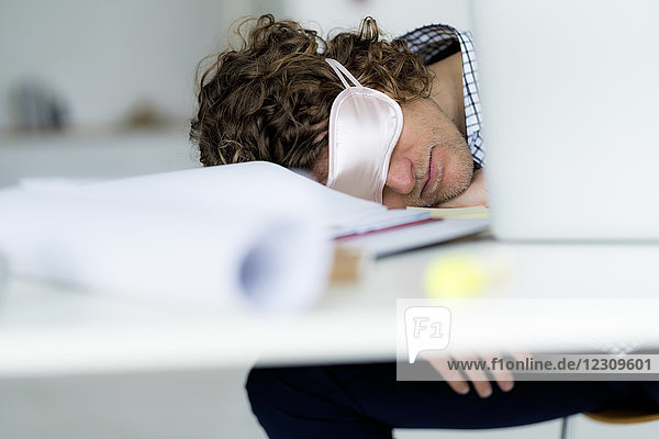 Überarbeiteter Geschäftsmann  der an seinem Schreibtisch schläft und eine Schlafmaske trägt.