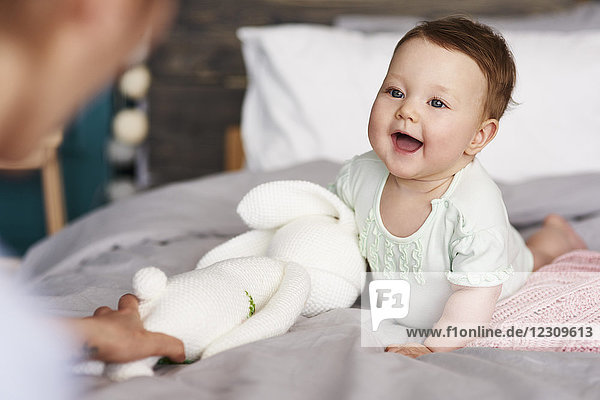 Glückliches Baby mit Kuscheltier auf dem Bett zu Hause mit Blick auf ihre Mutter