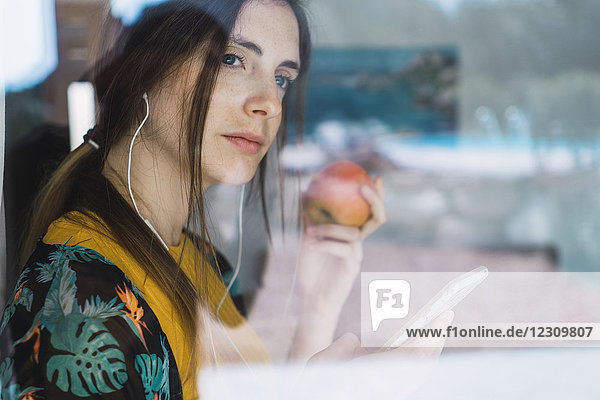 Junge Frau mit Kopfhörer  Handy und Apfel aus dem Fenster schauend
