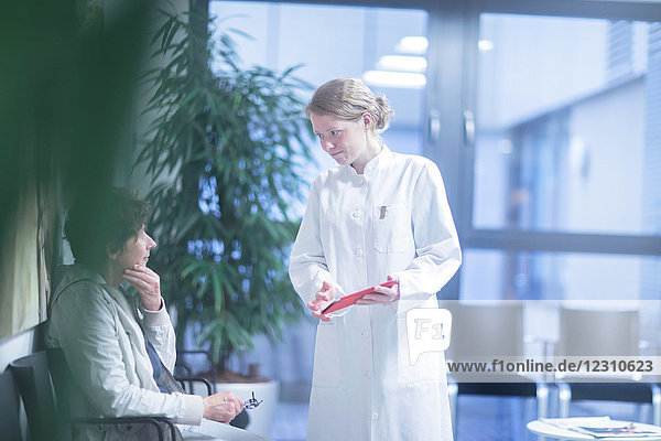 Radiologe  hält digitales Tablett in der Hand und spricht mit reifer Frau im Wartebereich des Krankenhauses
