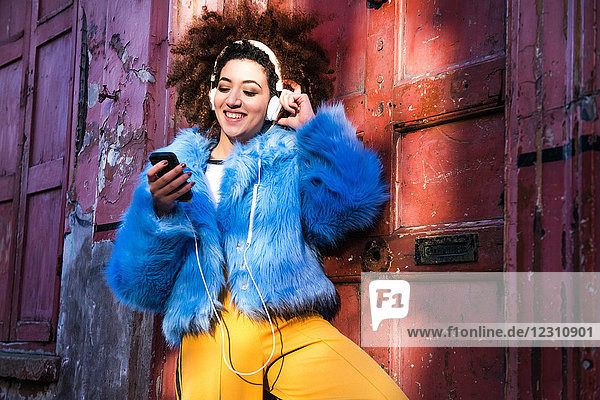 Portrait of woman with afro hair wearing fur coat  listening to music through headphones on smartphone