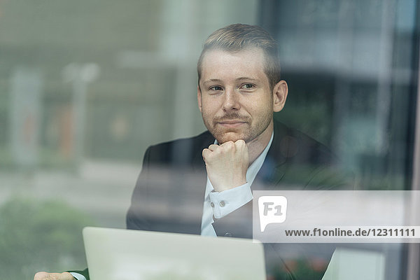 Businessman sitting  laptop in front of him  thoughtful expression  viewed through window