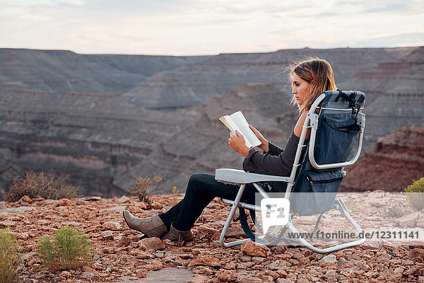 Junge Frau in abgelegener Umgebung  sitzt auf Campingstuhl  liest Buch  Mexican Hat  Utah  USA