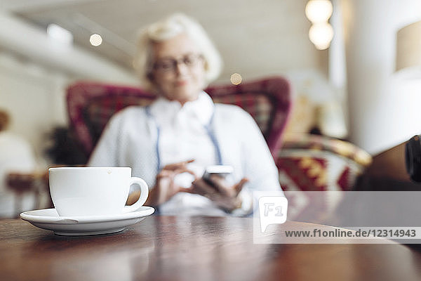 Coffee cup on table and senior woman using mobile phone during coffee break in cafe