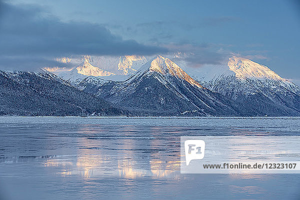 Das warme Licht des Sonnenuntergangs beleuchtet die schneebedeckten Gipfel des Turnagain Passes  die Berge spiegeln sich auf der eisigen Meeresoberfläche  Seward Highway  Süd-Zentral-Alaska im Winter; Alaska  Vereinigte Staaten von Amerika