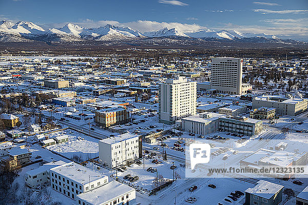Luftaufnahme der schneebedeckten 3rd und 4th Avenue in der Innenstadt von Anchorage  das Sheraton-Hotelgebäude erhebt sich mit den Chugach Mountains im Hintergrund im Winter  Süd-Zentral-Alaska; Anchorage  Alaska  Vereinigte Staaten von Amerika