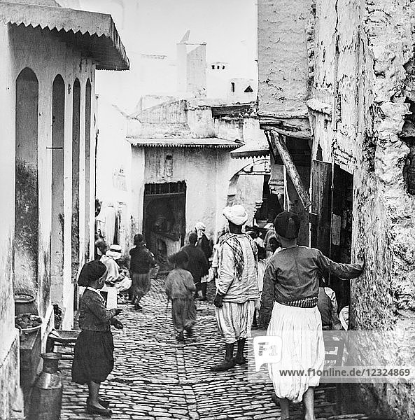 A narrow street with a cafe and pedestrians  Magic lantern slide  circa 1900; Algiers  Algeria