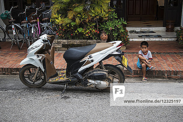 Boy sitting along Sisavangvong Road next to a parked motorcycle; Luang Prabang  Luang Prabang  Laos
