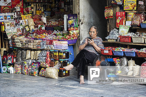 Verkäuferin sitzt barfuß vor ihrem Laden und benutzt ihr Smartphone; Hanoi  Hanoi  Vietnam
