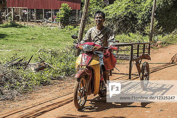 Ein Mann fährt auf einem Motorrad und zieht einen Karren über eine unbefestigte Straße; Siem Reap  Kambodscha