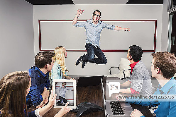 Ein junger Geschäftsmann springt  um eine Leistung mit seinen Mitarbeitern zu feiern  die während einer Präsentation in einem Konferenzraum arbeiten  während einer seiner Mitarbeiter ein Foto mit einem Pad macht; Sherwood Park  Alberta  Kanada