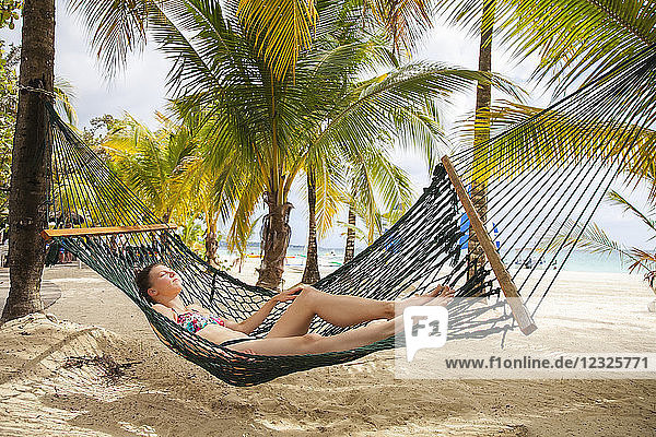 A young woman in a bikini lays in a hammock on a tropical beach with the ocean in the background; Negril Jamaica