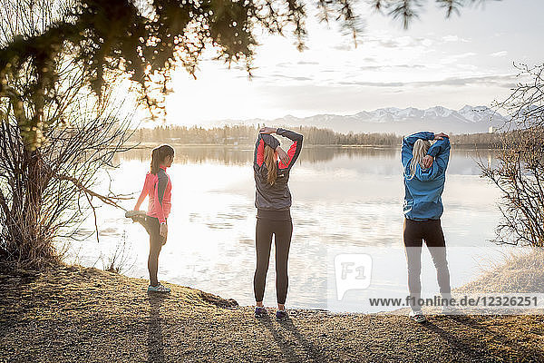 Three young women stretching on a trail at the water's edge; Anchorage  Alaska  United States of America