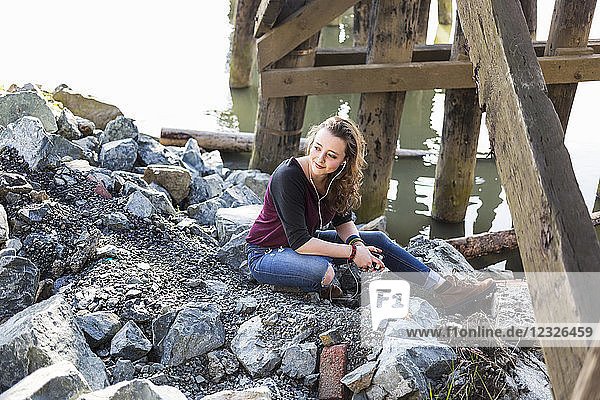 Porträt einer jungen Frau  die auf Felsen unter einer Brücke am Wasser sitzt und Musik hört
