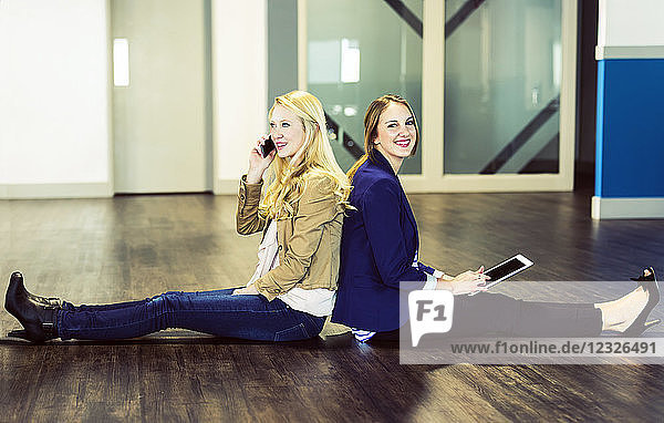Zwei hübsche junge Frauen  die als Geschäftsleute der Jahrtausendwende Rücken an Rücken auf dem Boden ihres Arbeitsplatzes sitzen und Technologie nutzen; Sherwood Park  Alberta  Kanada
