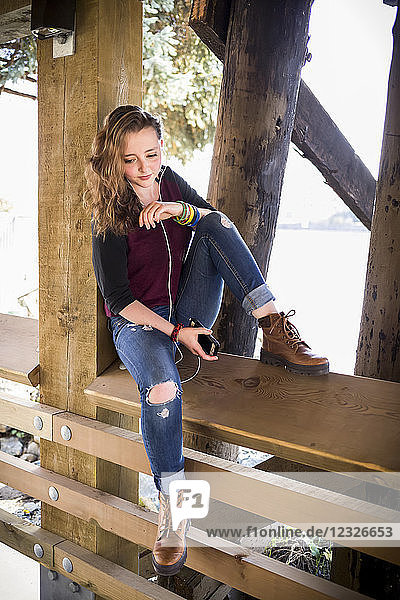 Eine junge Frau sitzt auf einem Holzgeländer unter einer Brücke am Wasser und hört Musik; New Westminster  British Columbia  Kanada