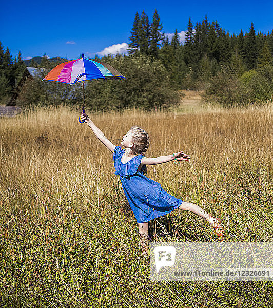 A young girl stands in a grass field holding an umbrella high and balancing on one foot; Salmon Arm  British Columbia  Canada