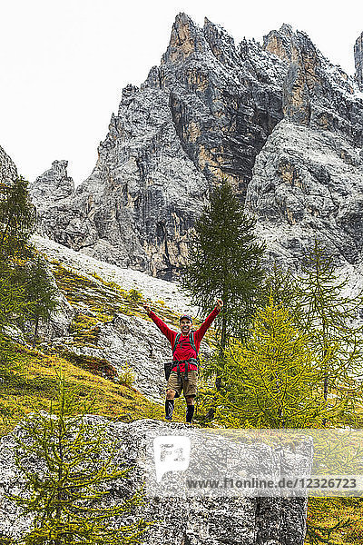 Männlicher Wanderer mit erhobenen Armen auf einem großen Felsen mit felsigem Abhang und Berggipfel; Sexten  Bozen  Italien